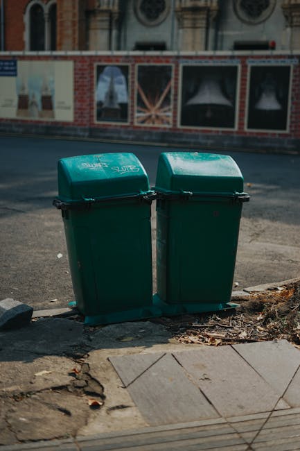 A street cleaning worker wearing an orange high-visibility uniform and a matching cap, using a long-handled tool to sweep debris from the curb into a green collection cart filled with trash and litter